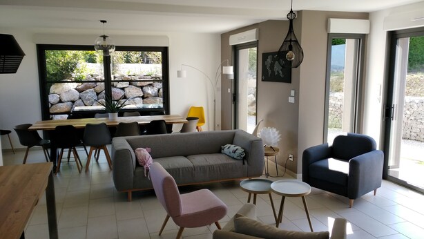 Ein modernes Wohnzimmer mit einem grauen Sofa, einem pinken Sessel, einem blauen Sessel und einem Holzespeisetisch mit Stühlen. Ein großes Fenster bietet Blick auf Felsen und Grünflächen.