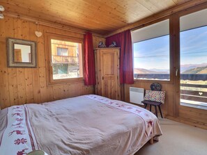 A wooden cabin room with a bed, a window with curtains, a chair, and a view of mountains.