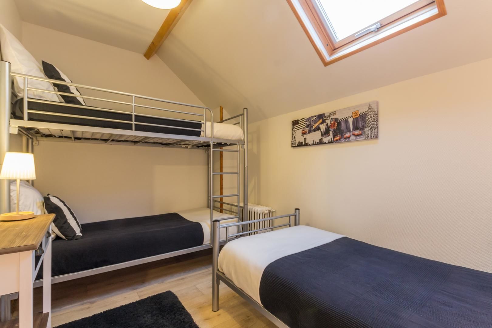 A dormitory room with bunk beds, a desk with a lamp, a radiator, and a skylight.