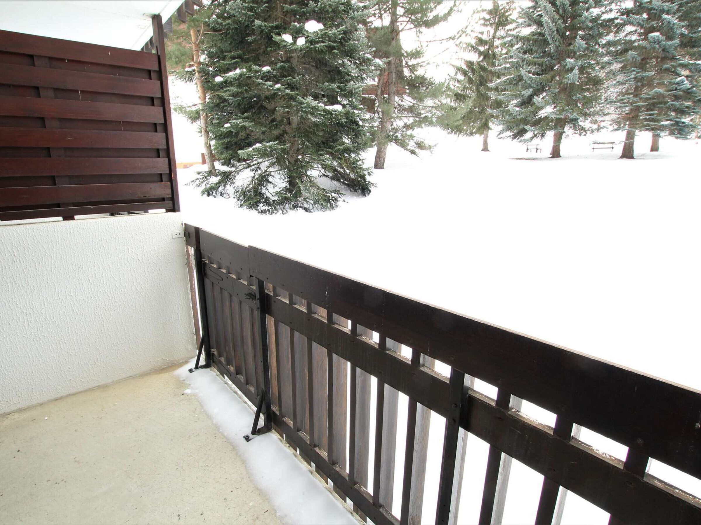 A balcony with a wooden railing, snow on the ground, and evergreen trees in the background.
