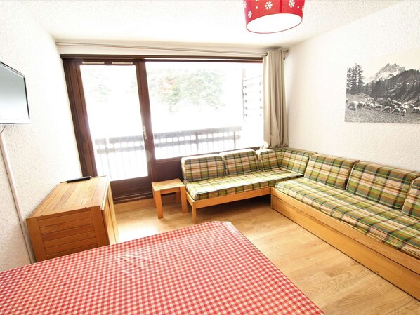 Um quarto com uma mesa xadrez, um sofá de retalhos, um armário de madeira e uma janela com vista para uma varanda.
