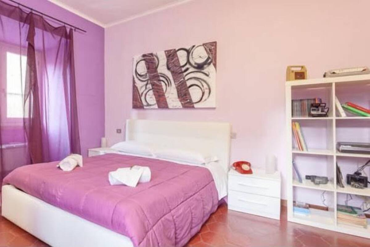 Un dormitorio con cama, mesita de noche, estantería y ventana con cortinas translúcidas.