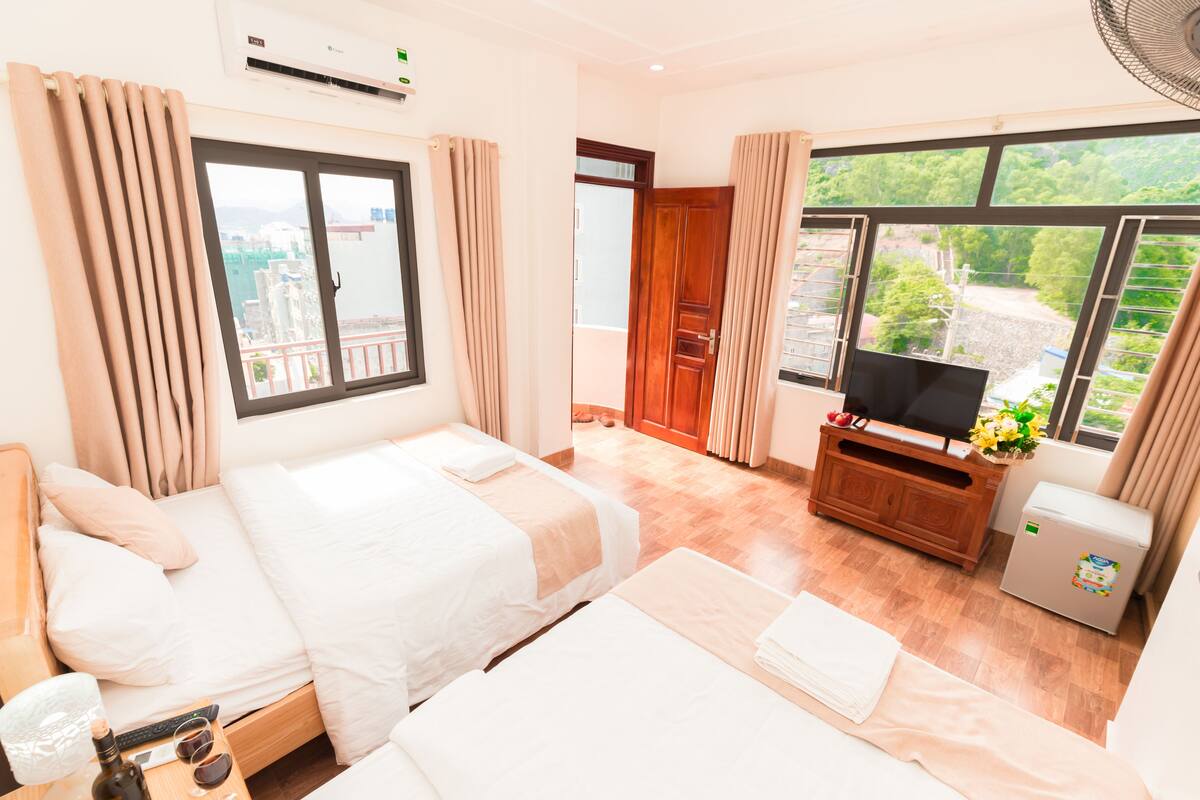 A hotel room with two beds, a TV, a mini-fridge, and a balcony with a view of greenery.