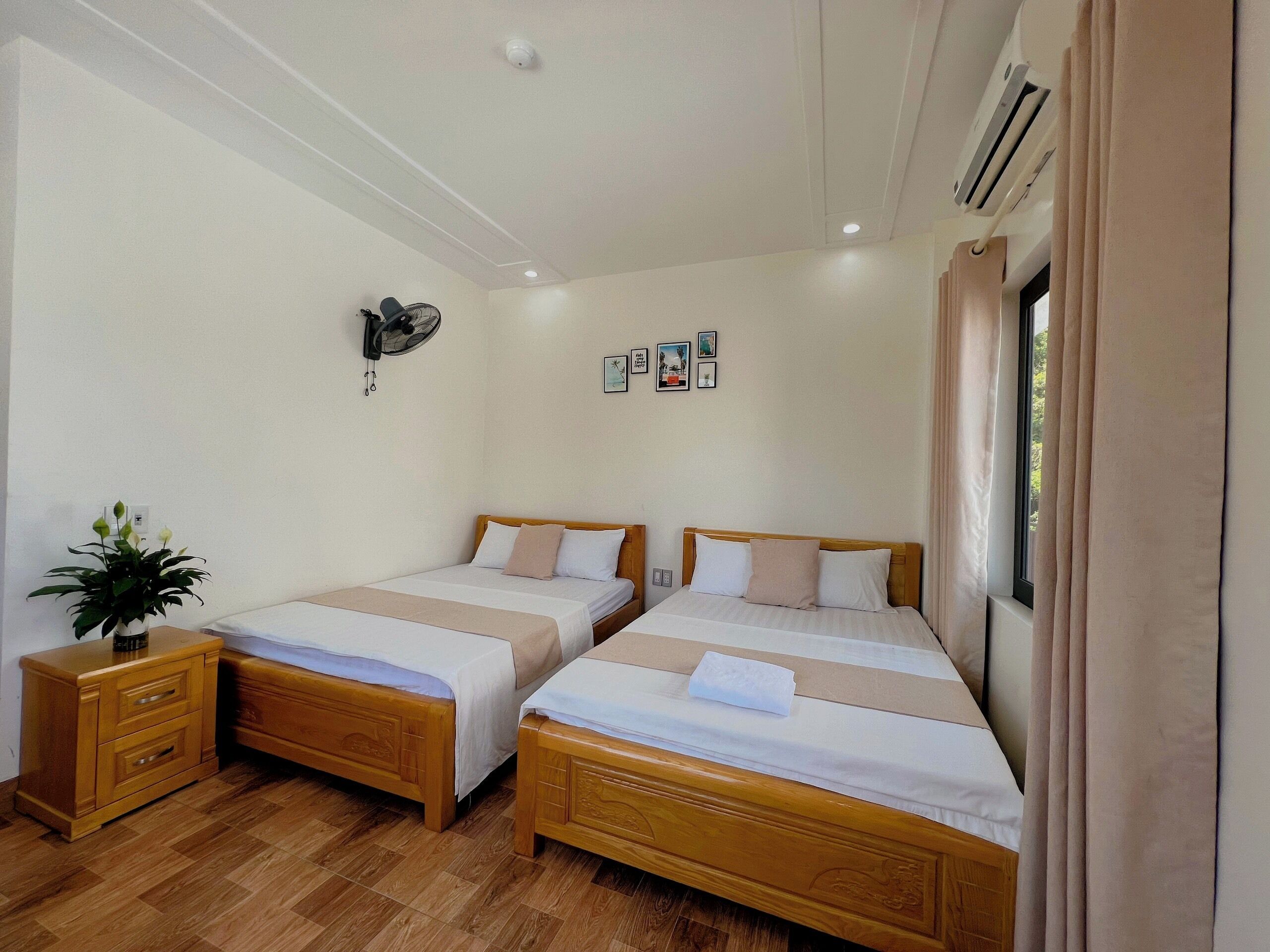 A hotel room with two wooden beds, a small bedside table with a plant, and a wall-mounted air conditioner.