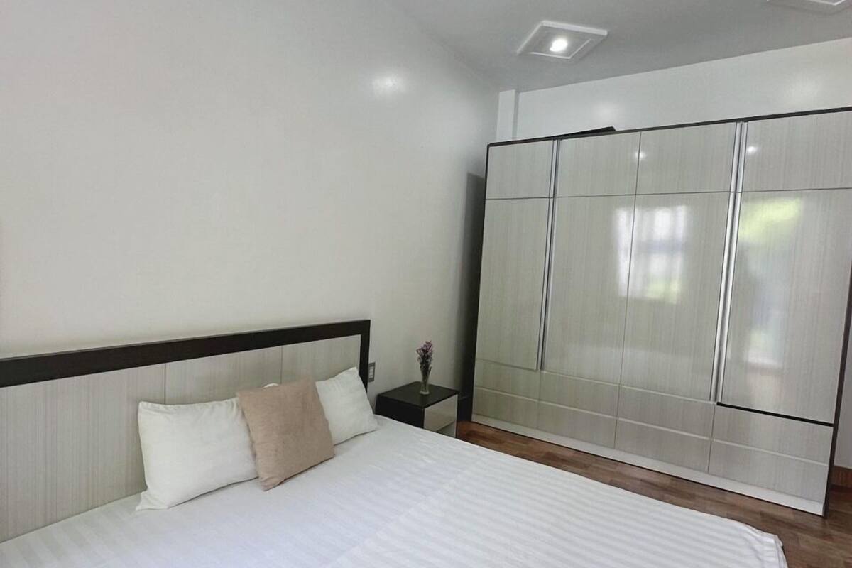 A neatly made bed with white linens and a rolled towel, a nightstand with a vase, and a wardrobe with sliding doors.