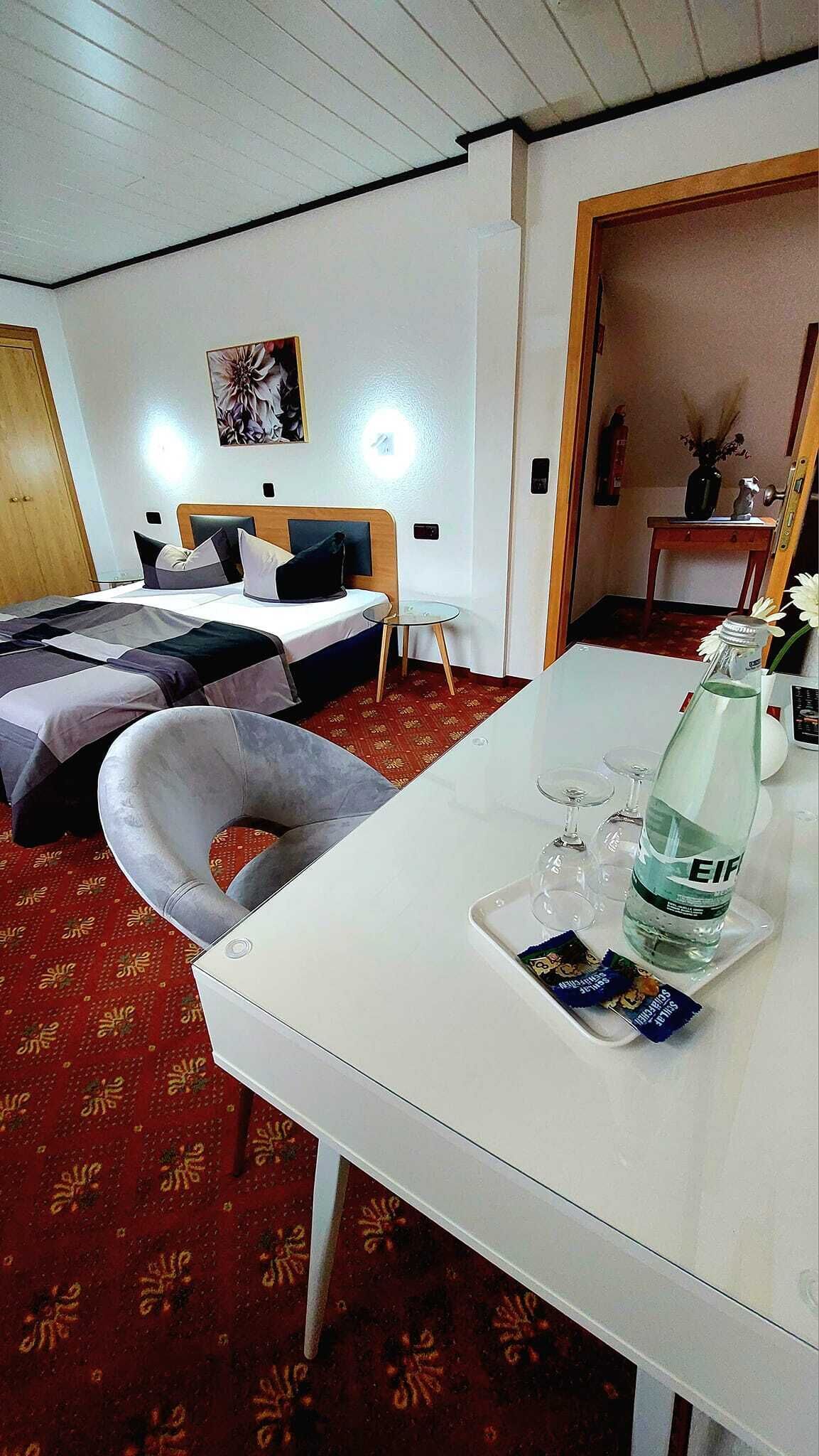 A hotel room with a bed, a small table with a bottle and snacks, a chair, and a red carpet.