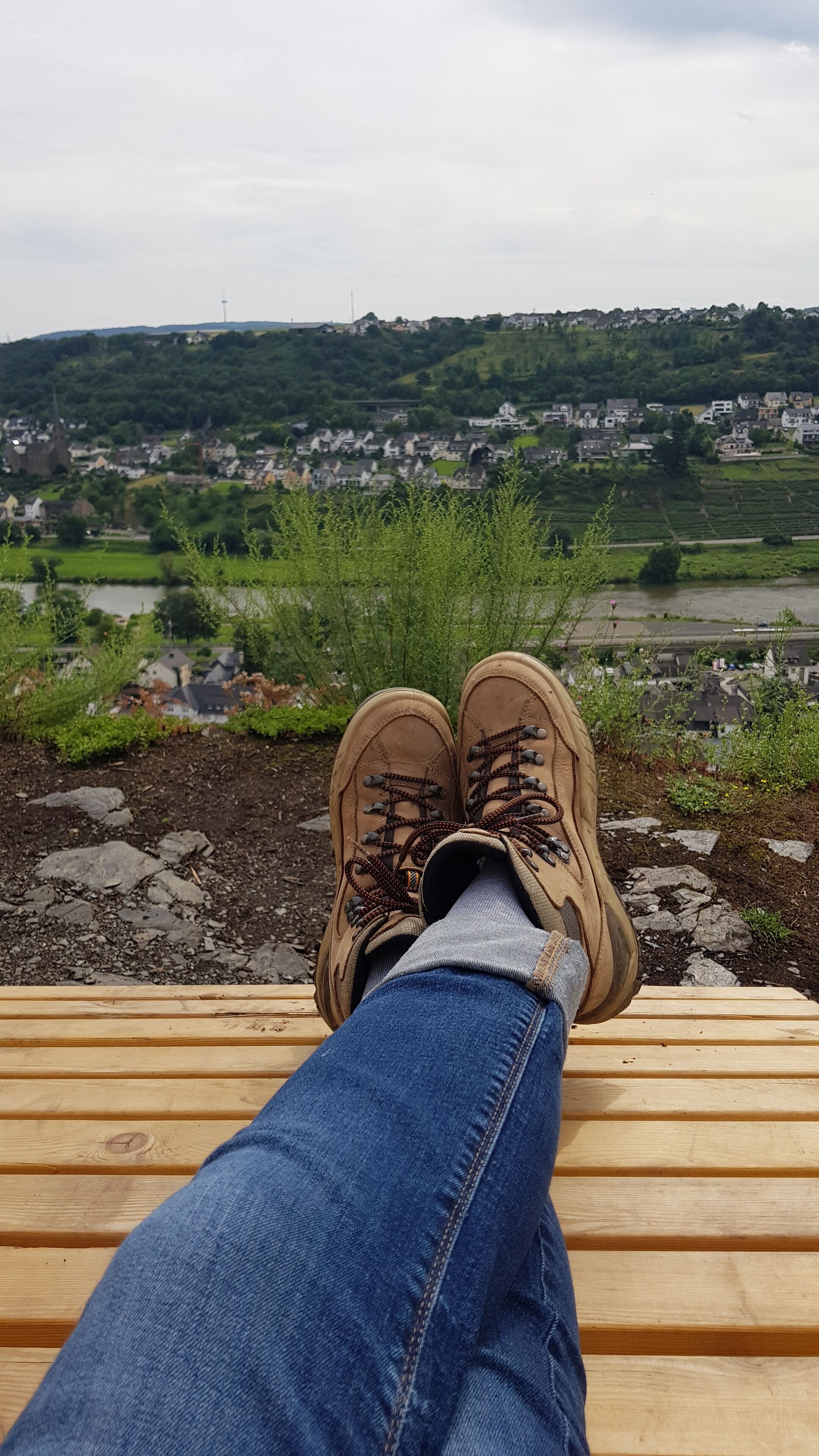 Un paio di scarponcini da trekking su una panca di legno con vista su un paesaggio rurale con case e campi.