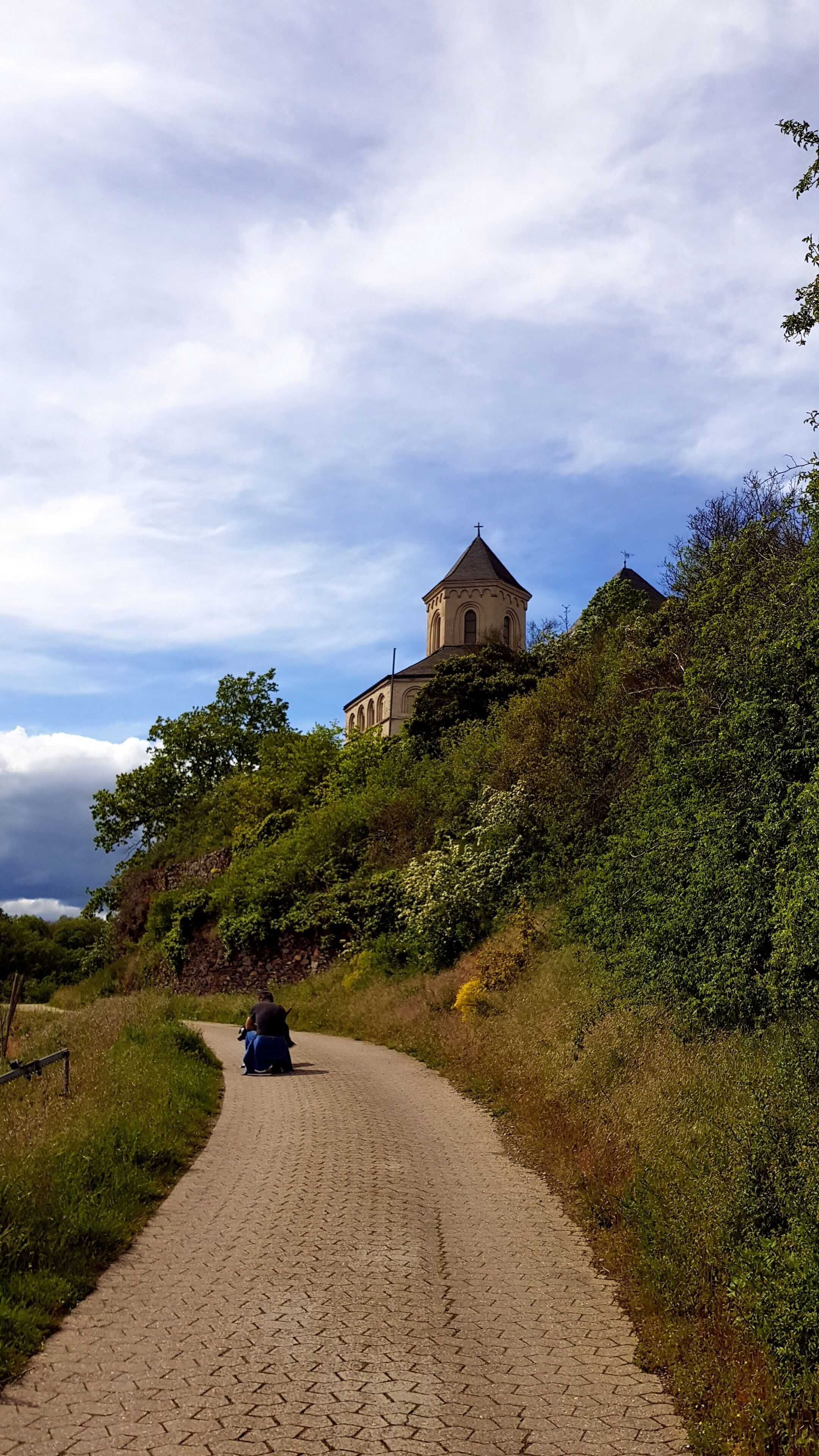 A paved pathway leading to a building with a cross on top, surrounded by greenery.