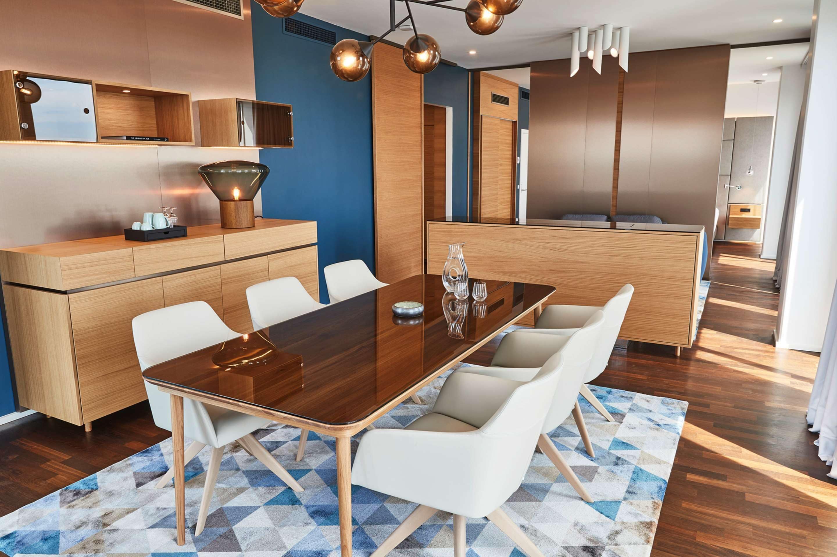 A modern dining area with a wooden table and white chairs, a kitchen in the background, and a blue accent wall.