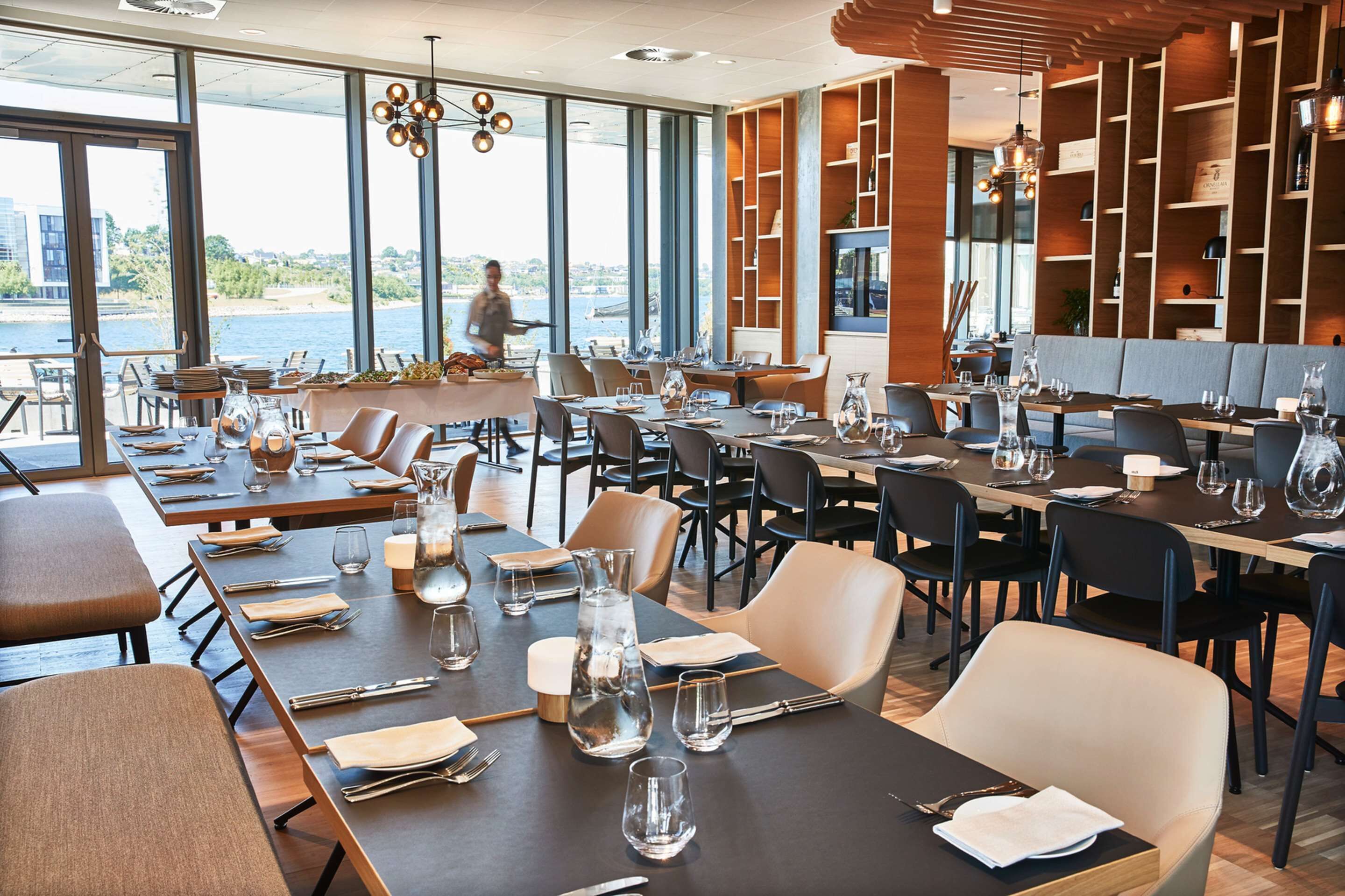 Un restaurante con mesas dispuestas para comer, grandes ventanales que ofrecen vistas al agua, y un diseño de interiores moderno.