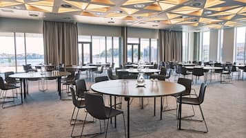 Un salón de conferencias con varias mesas y sillas, amplias ventanas con cortinas y un diseño geométrico en el techo.