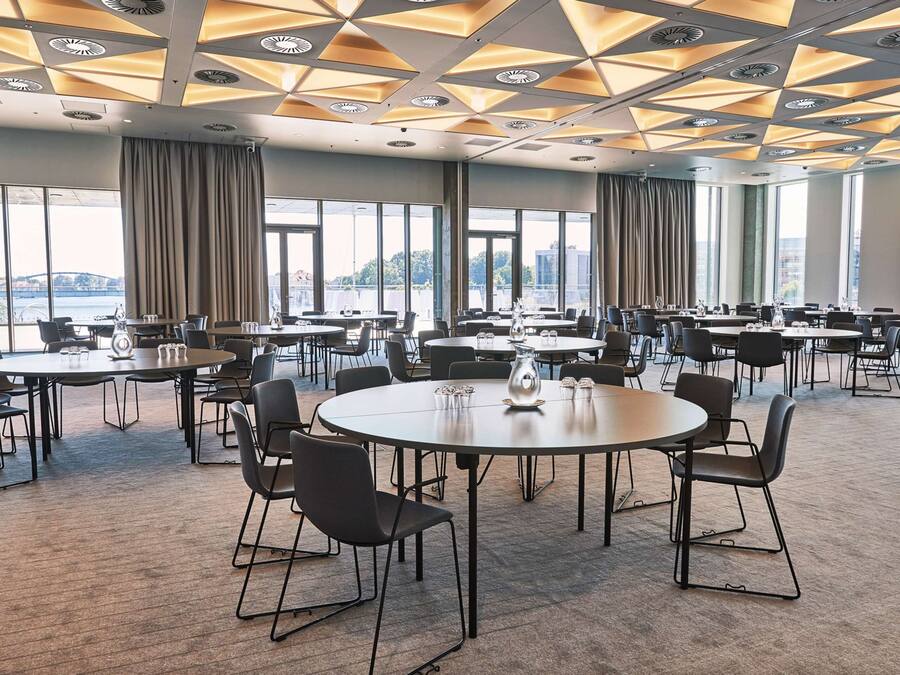 Un salón de conferencias con varias mesas y sillas, amplias ventanas con cortinas y un diseño geométrico en el techo.