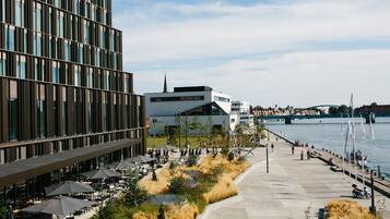 A riverside walkway with outdoor seating, a modern building, and a bridge in the background.