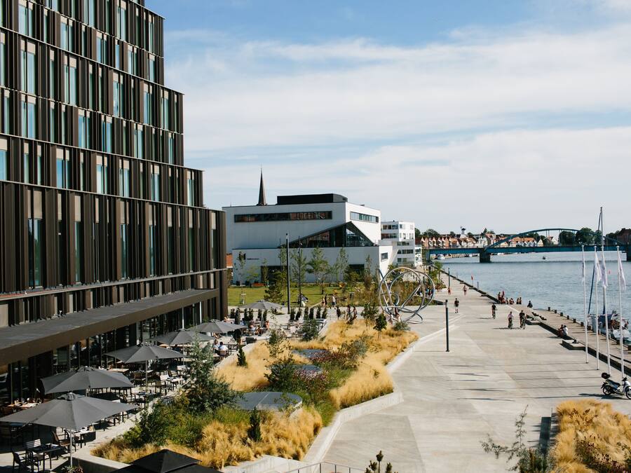 A riverside walkway with outdoor seating, a modern building, and a bridge in the background.