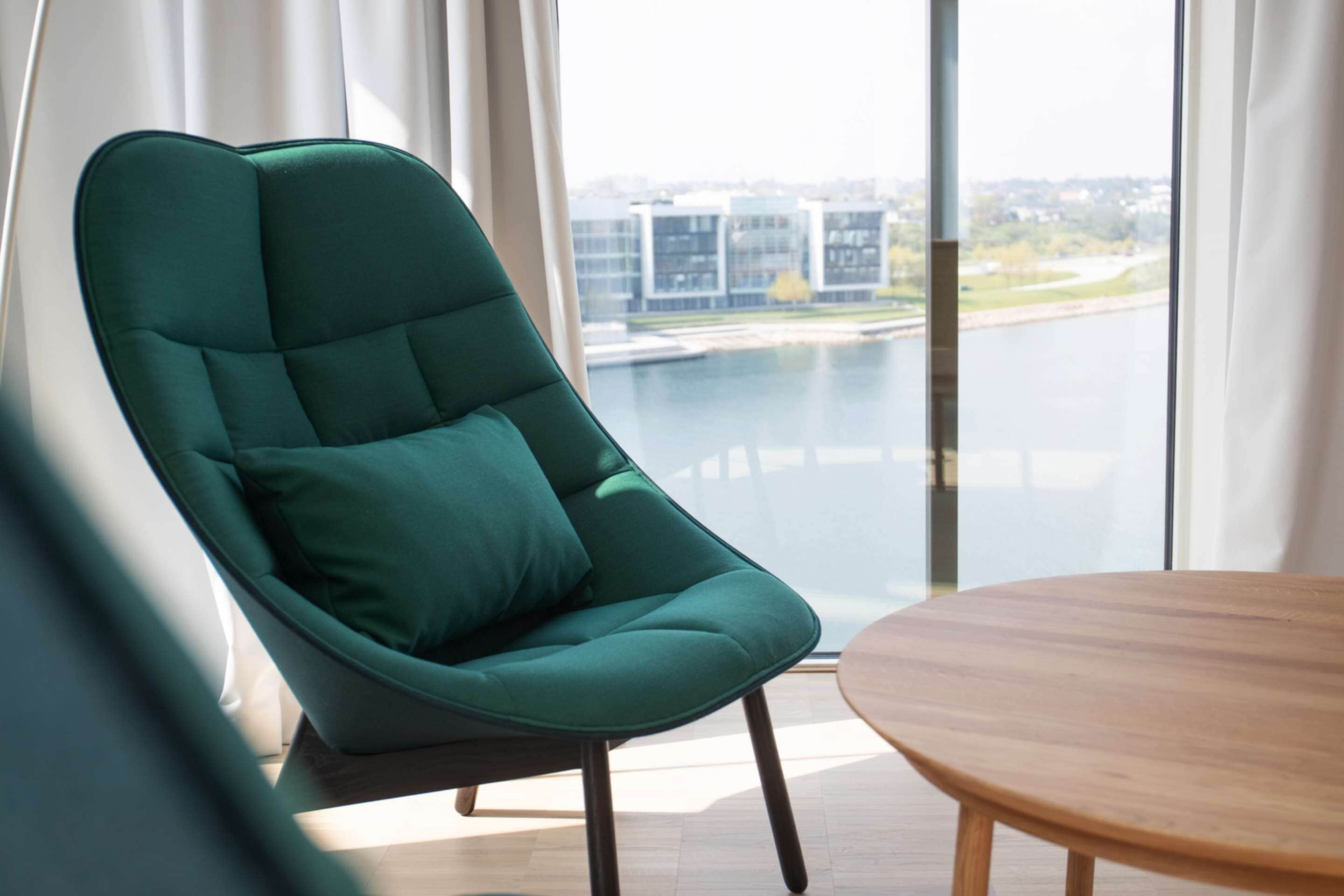 A modern room with a green upholstered chair, a wooden table, and a large window offering a view of buildings and water.