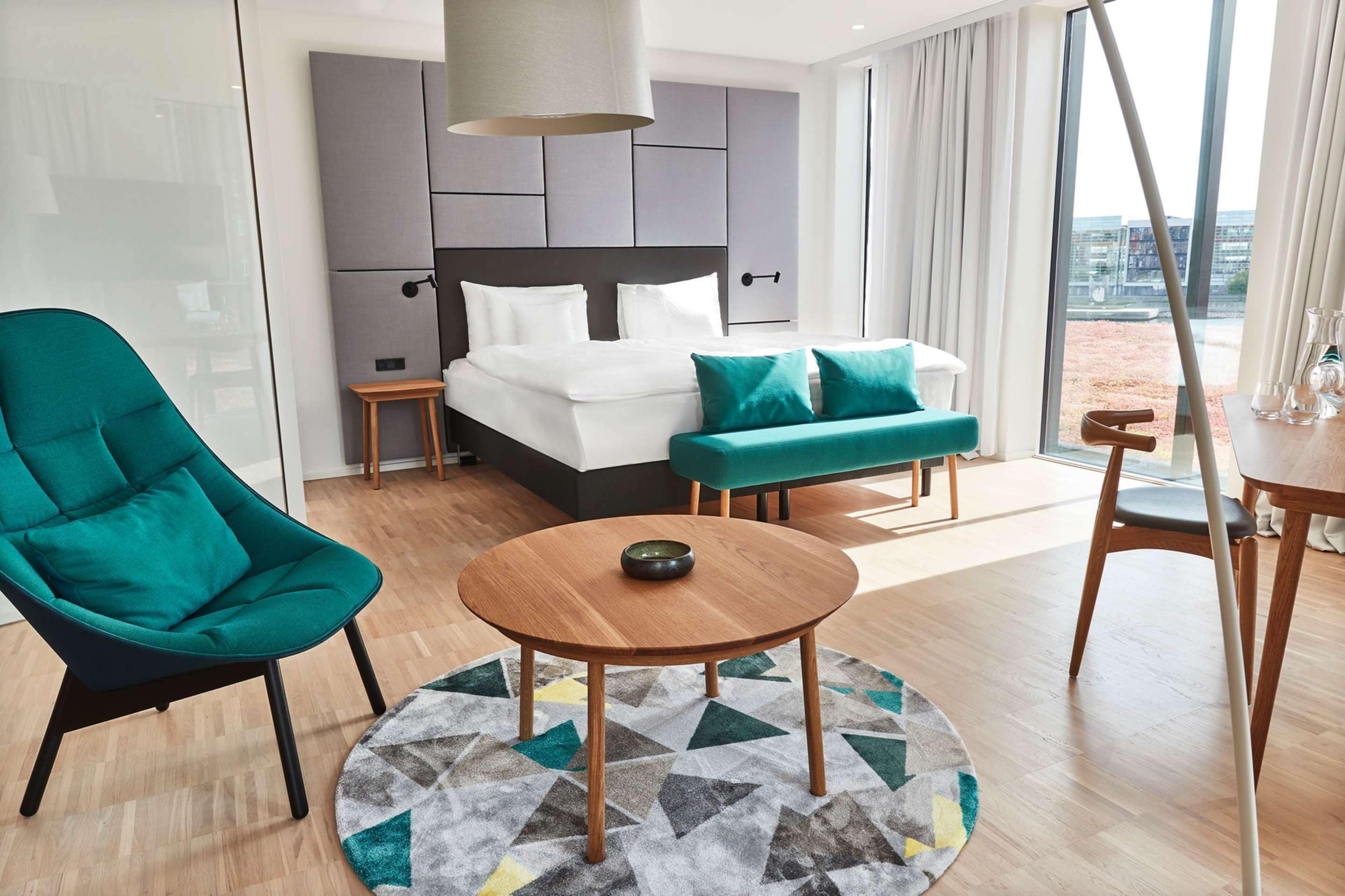 A modern hotel room with a large bed, a green armchair, a wooden coffee table, and a view of the city.