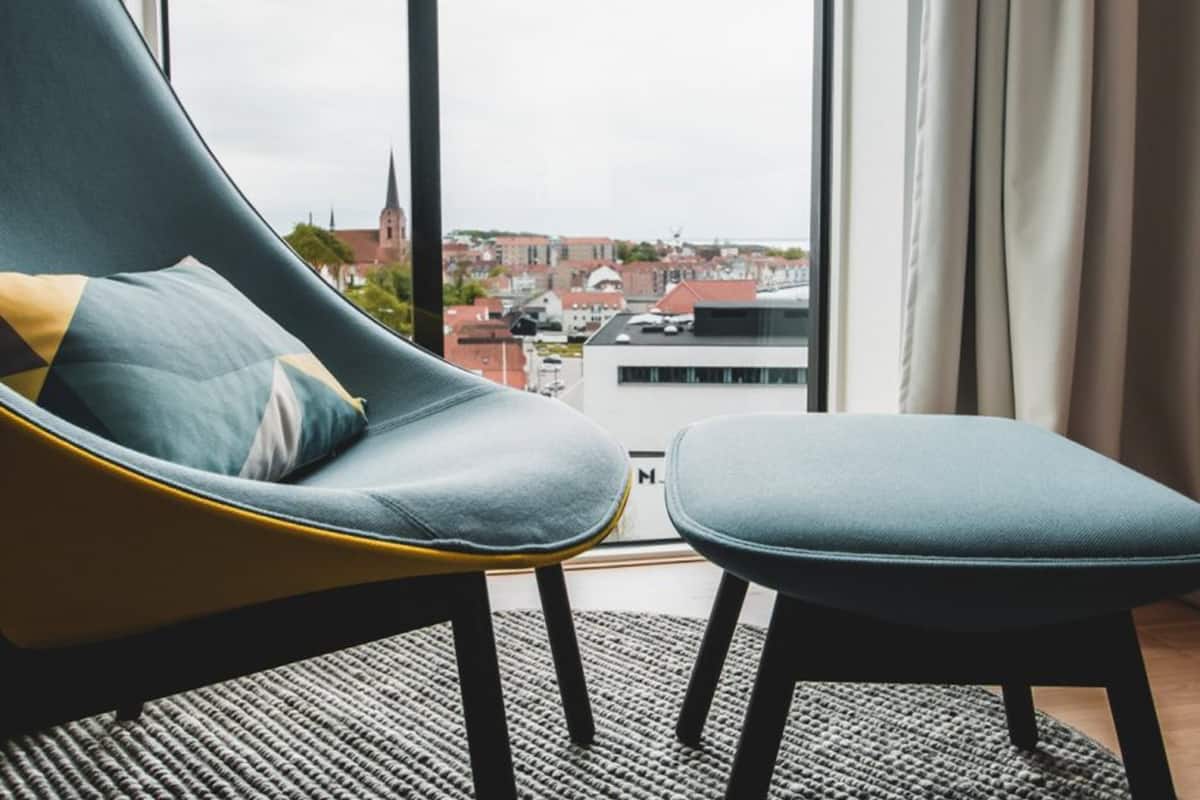 Une pièce moderne avec un fauteuil et un pouf au design incurvé, une grande fenêtre offrant une vue sur le paysage urbain, et un tapis noir et blanc.