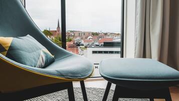Una habitación moderna con un sillón curvo y reposapiés, un ventanal con vistas a un paisaje urbano, y una alfombra blanco y negro.