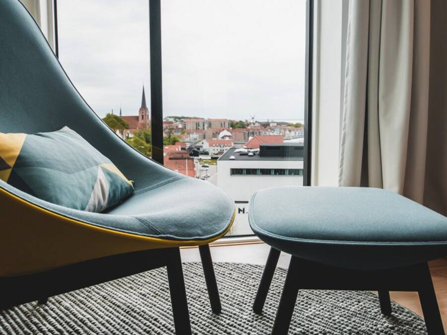 Una habitación moderna con un sillón curvo y reposapiés, un ventanal con vistas a un paisaje urbano, y una alfombra blanco y negro.