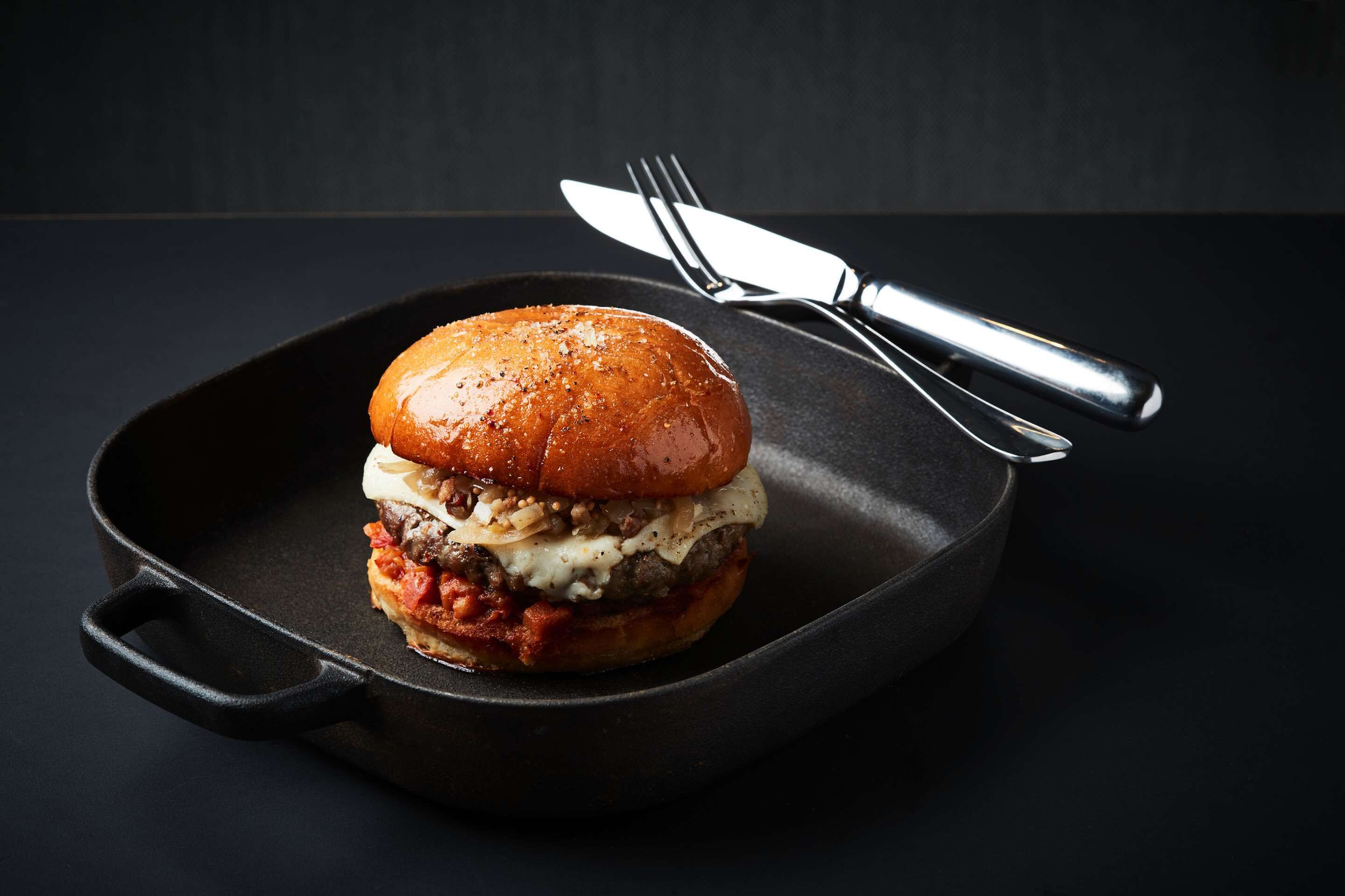 A burger in a cast iron skillet with a fork and knife beside it.
