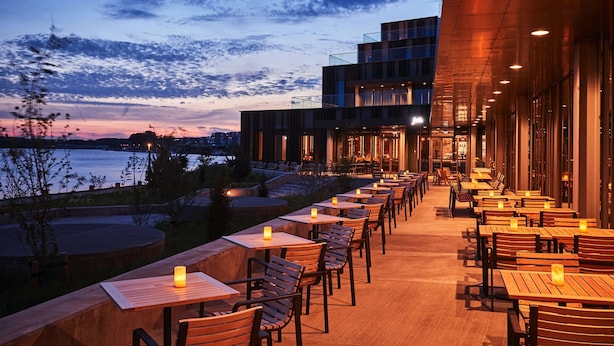 Un área de comedor al aire libre con mesas y sillas, con vistas a una masa de agua y un edificio moderno con ventanas de vidrio.