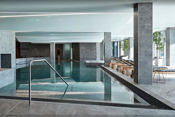Una piscina interior moderna con paredes de vidrio transparente, una barandilla de metal y suelo de piedra.