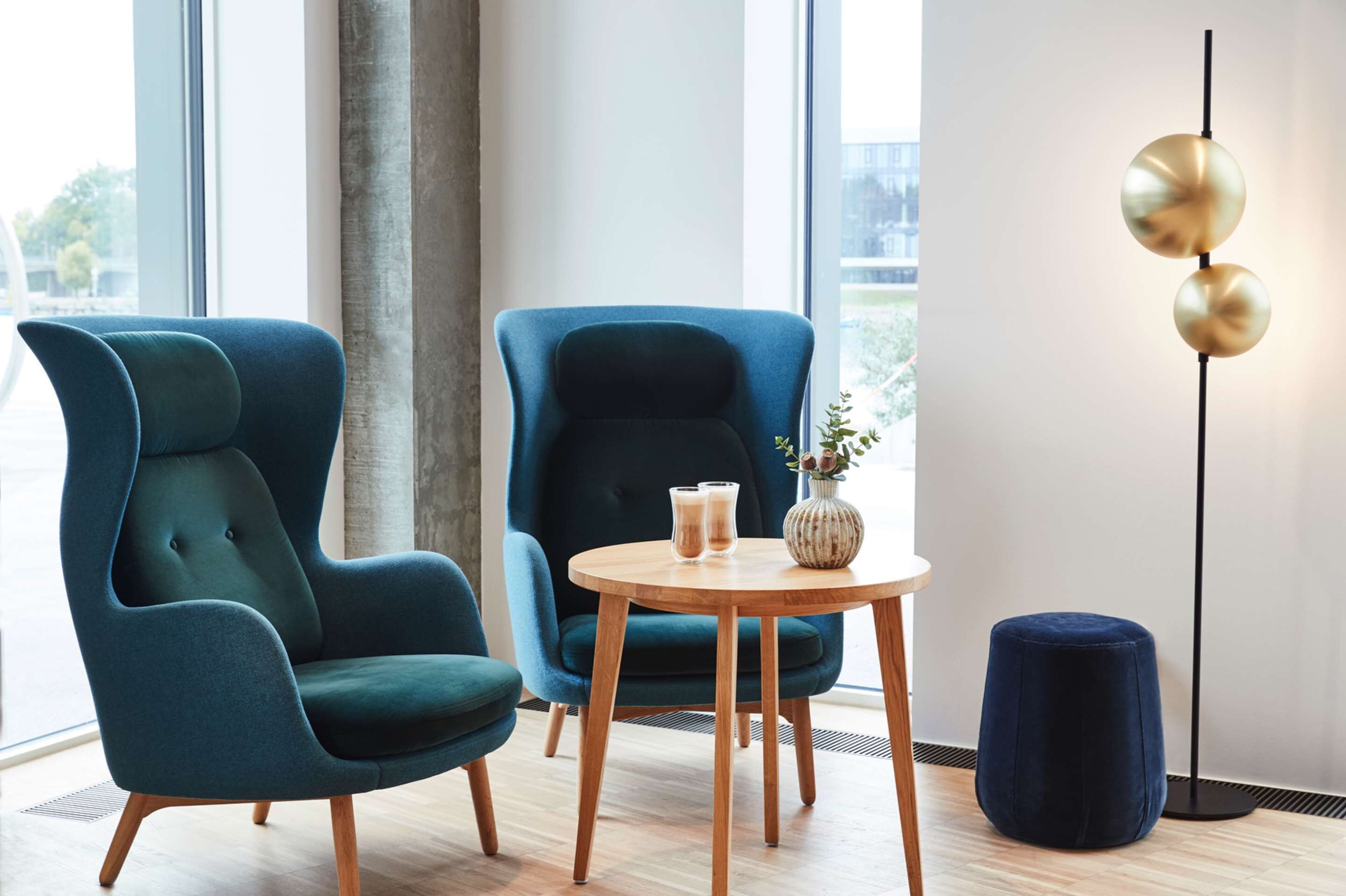 Ein modernes Zimmer mit zwei petrolfarbenen Sesseln, einem Holztisch mit Getränken, einer Vase mit Blumen und einer stehenden Stehlampe.