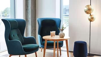 Ein modernes Zimmer mit zwei petrolfarbenen Sesseln, einem Holztisch mit Getränken, einer Vase mit Blumen und einer stehenden Stehlampe.