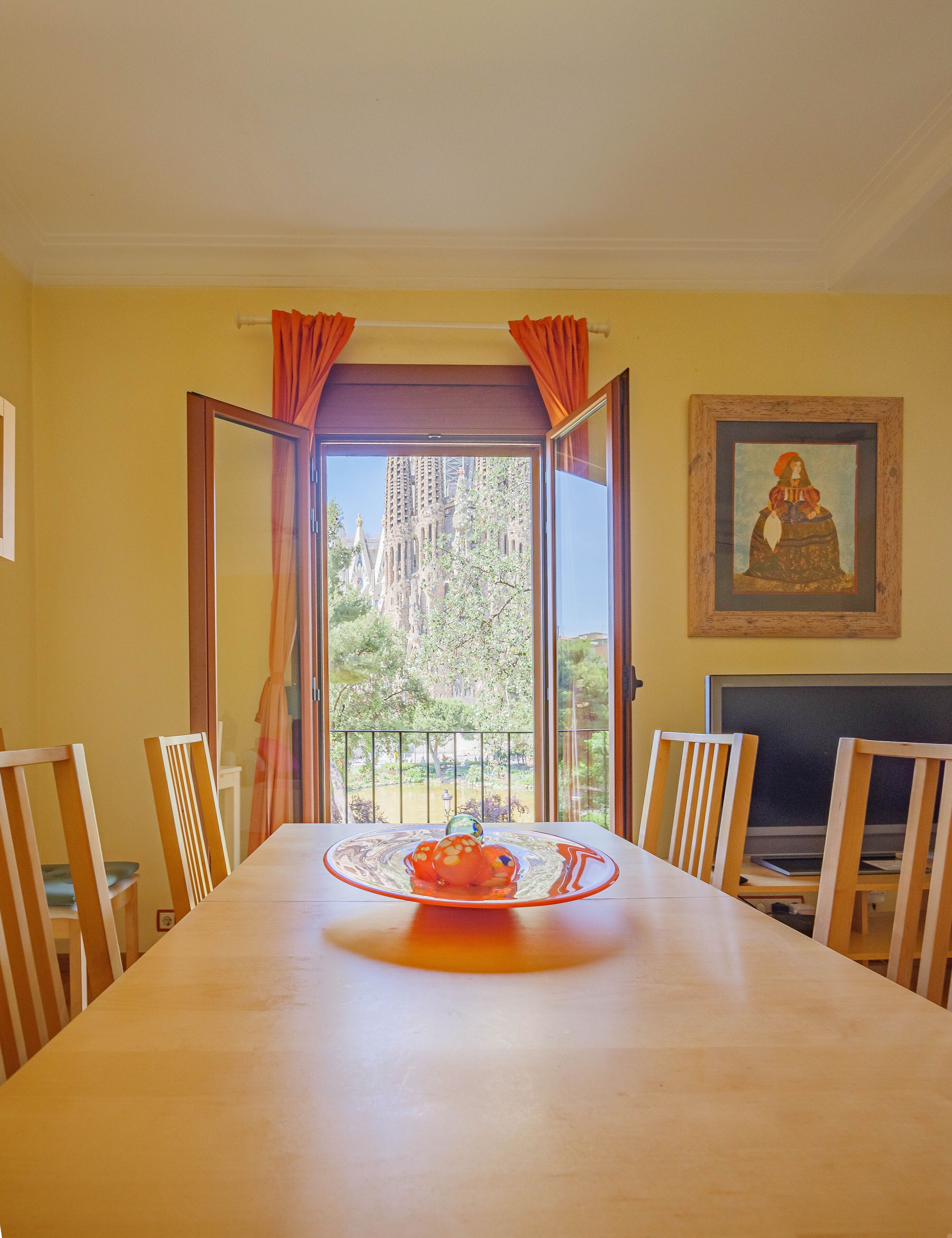 A dining area with a long table, chairs, a painting on the wall, and a view of tall buildings through an open door.