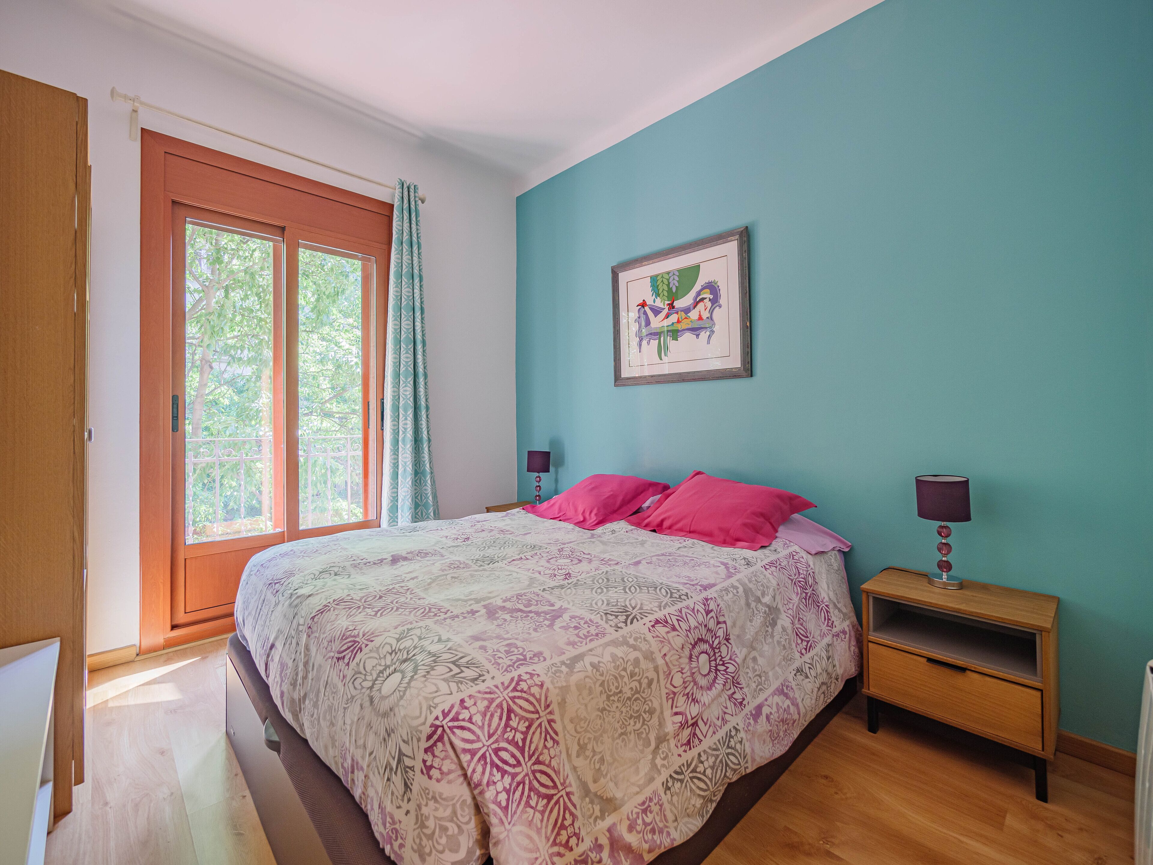 A bedroom with a bed, a nightstand, a wall-mounted lamp, a window with curtains, and a framed picture on the wall.