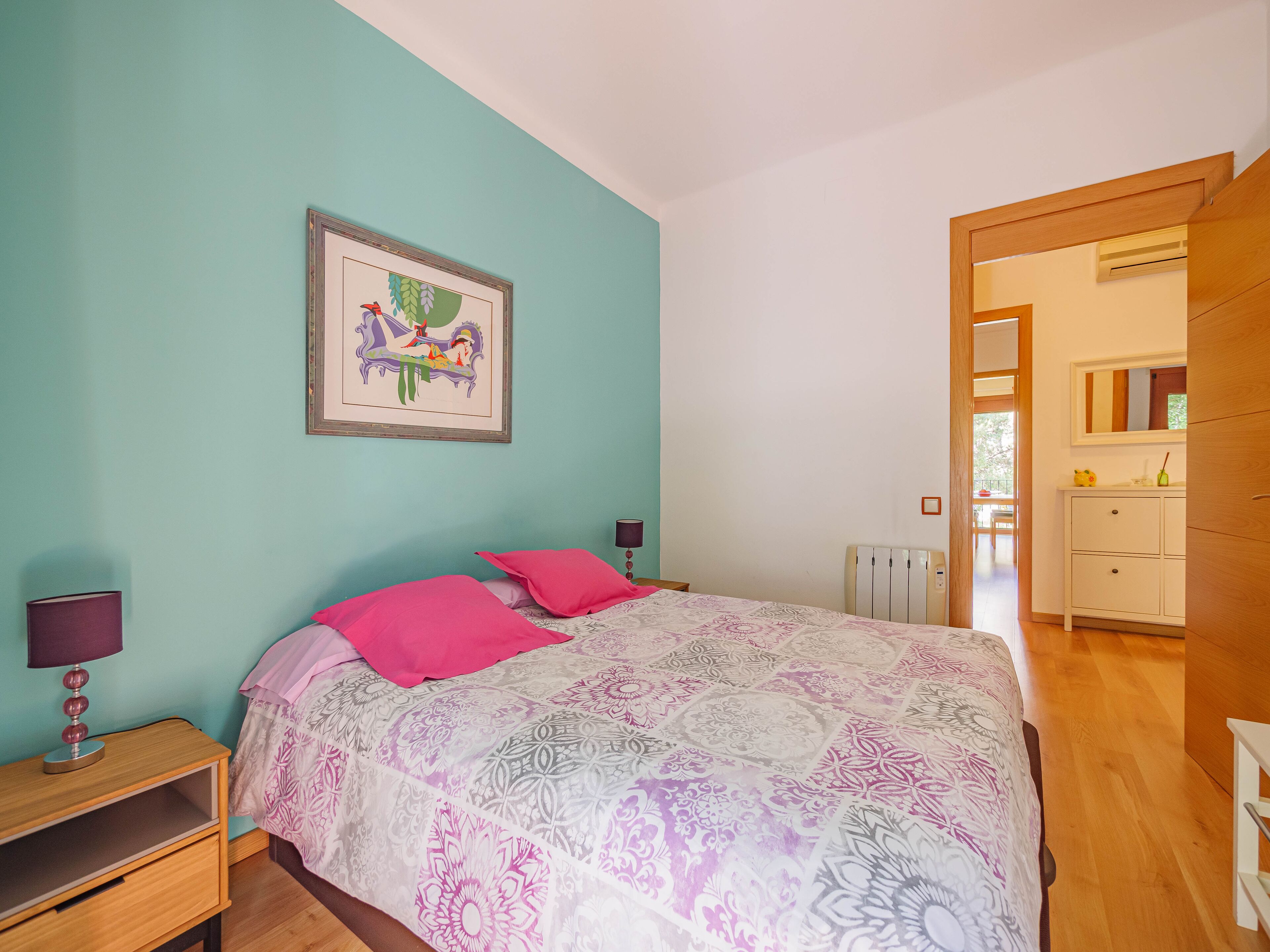 Un dormitorio con cama, mesita de noche, lámpara, un cuadro en la pared y vista al baño por una puerta abierta.