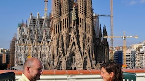 Two people sitting at a table with a bottle of champagne and a corkscrew, with a view of a cathedral under construction in the background. - Absolute Sagrada Familia (Barcelona)