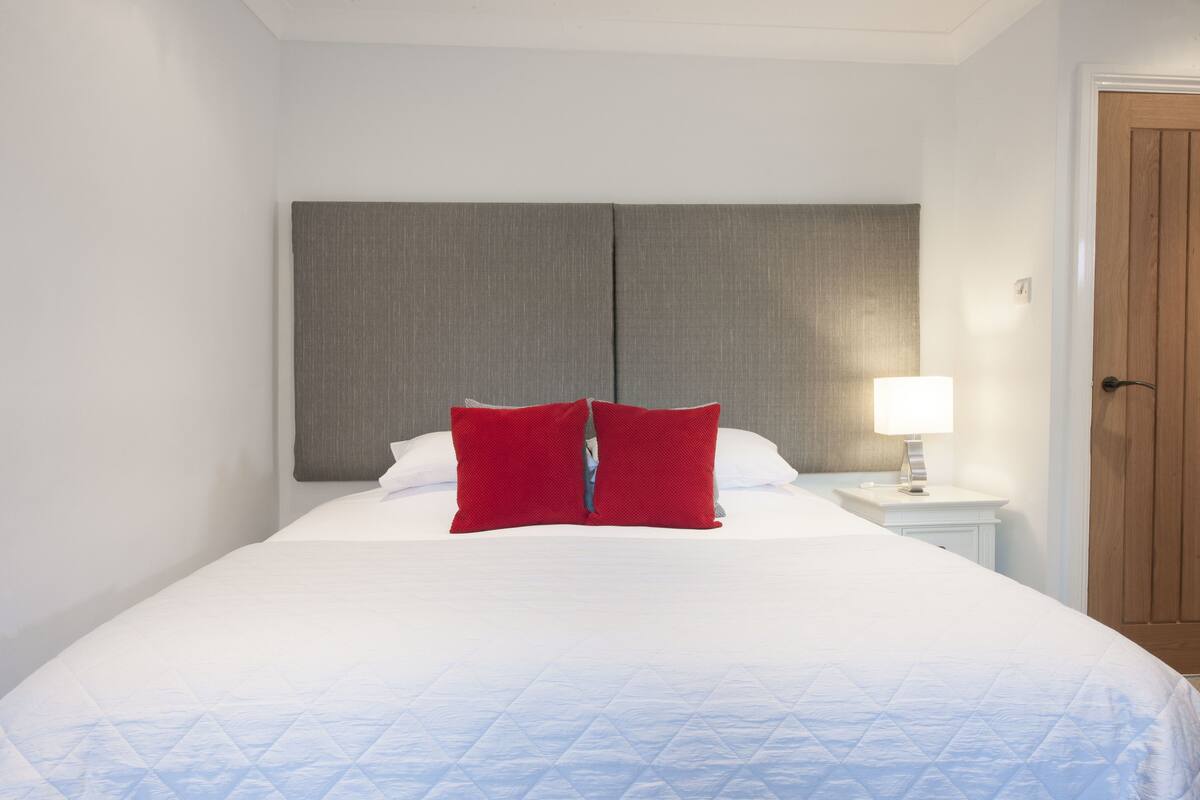 A neatly made bed with a white duvet and red pillows, a bedside lamp, and a wooden door.