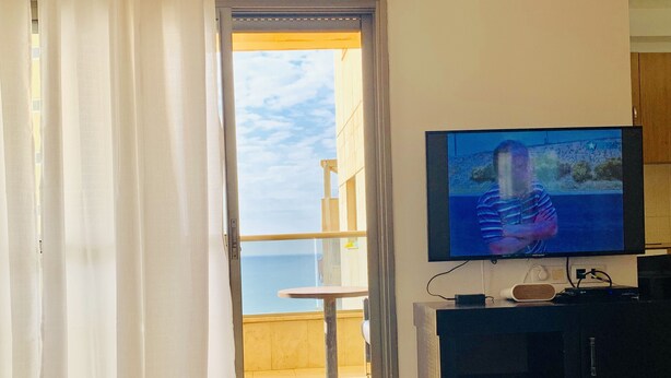 A room with a television on a stand, a small table, and a view of the sea through a balcony door.