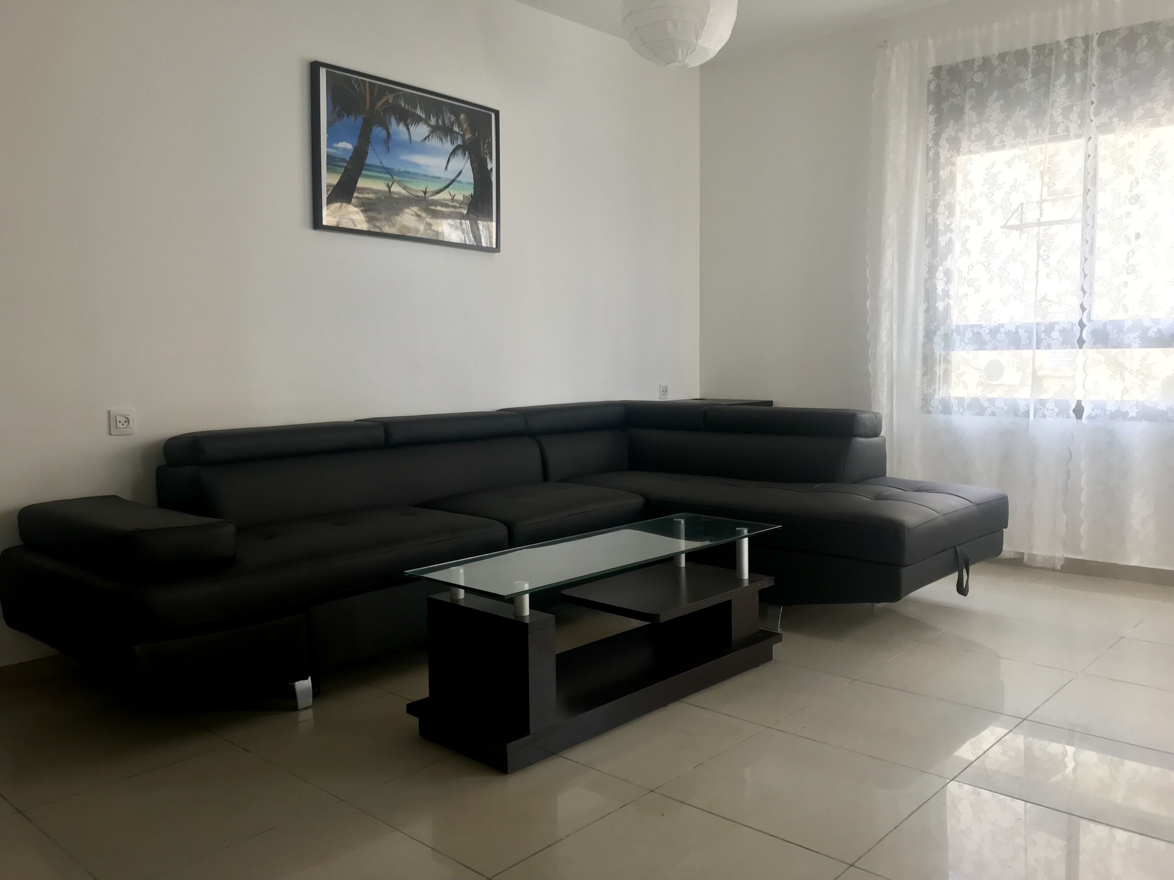 A modern living room with a black sectional sofa, a glass coffee table, and a beach-themed painting on the wall.