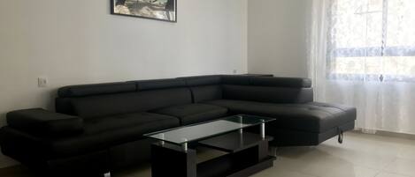 A modern living room with a black sectional sofa, a glass coffee table, and a beach-themed painting on the wall.