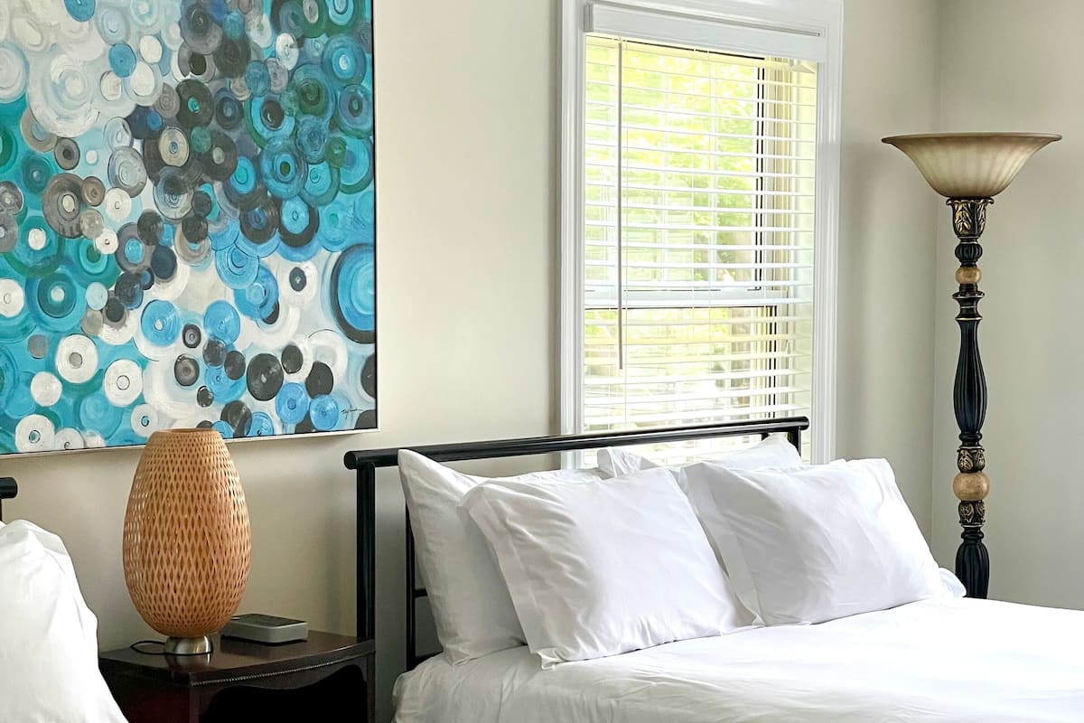 A hotel room with two beds, a large abstract painting, a window with blinds, and a tall floor lamp.