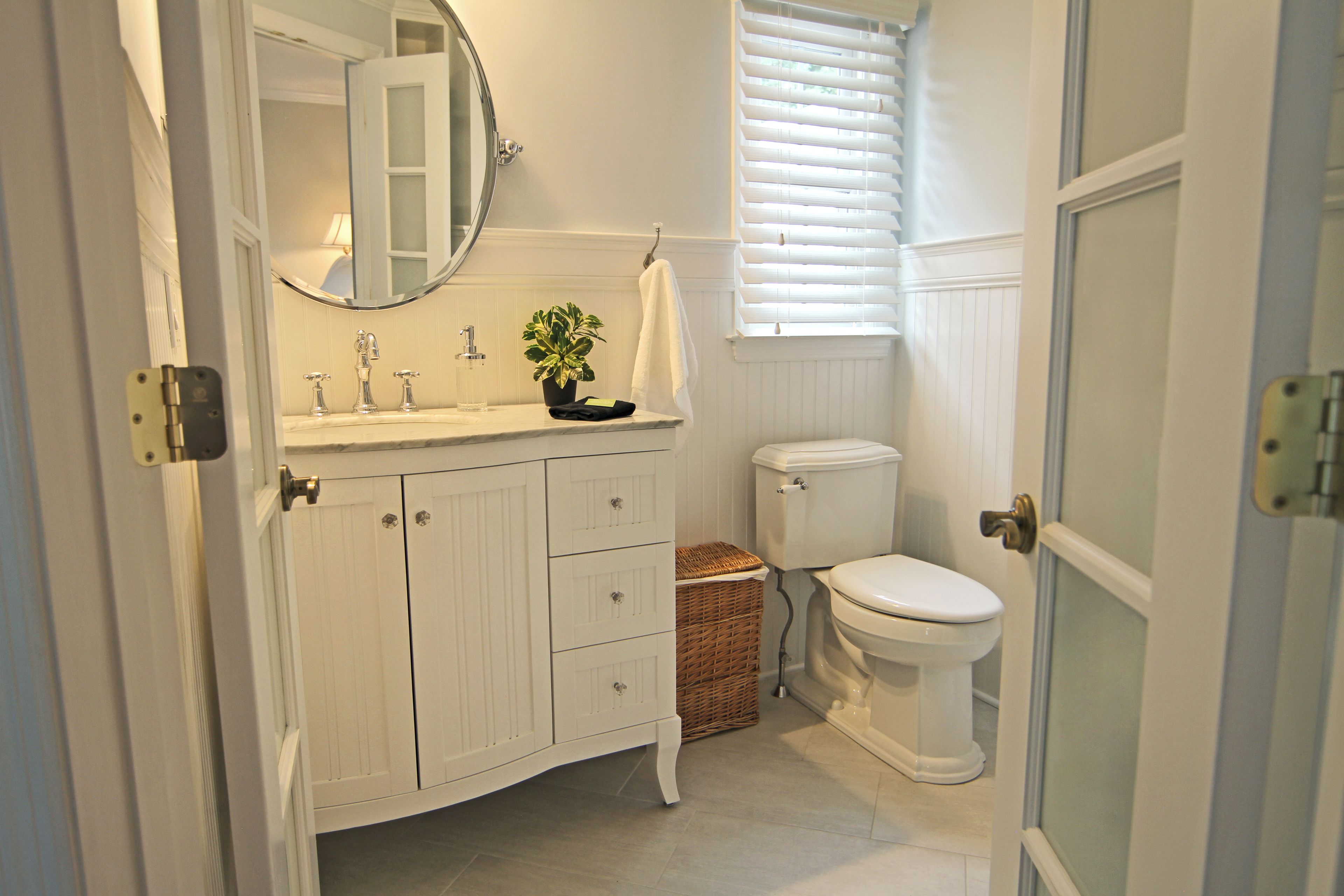 A bathroom with a white vanity, a round mirror, a toilet, and a window with blinds.