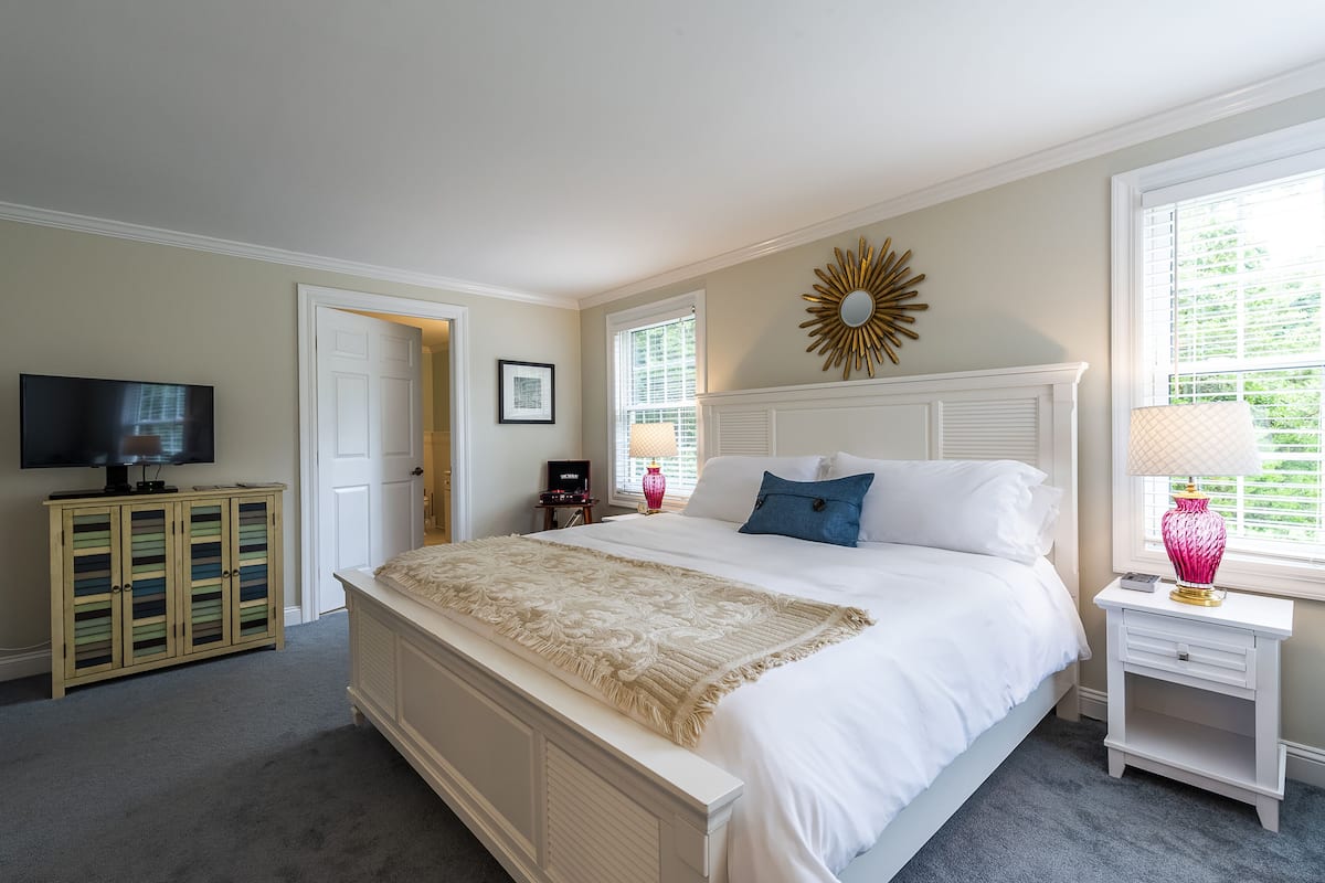 A bedroom with a large bed, a television on a stand, a dresser, and a window with blinds.