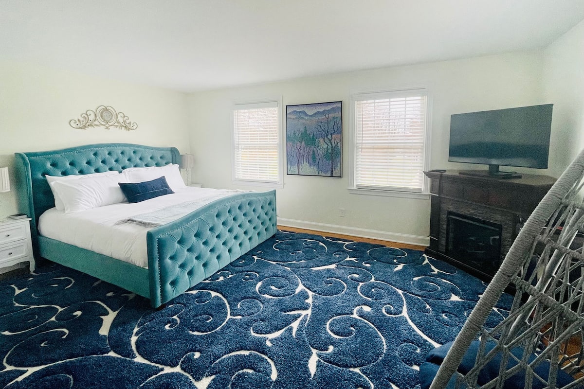 A bedroom with a teal upholstered bed, a patterned blue carpet, a television on a stand, and a fireplace.
