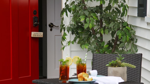Una puerta roja con un letrero que dice 'Fuera a la Playa', una silla de mimbre, una mesa con un plato de comida y bebidas, y una planta en maceta.
