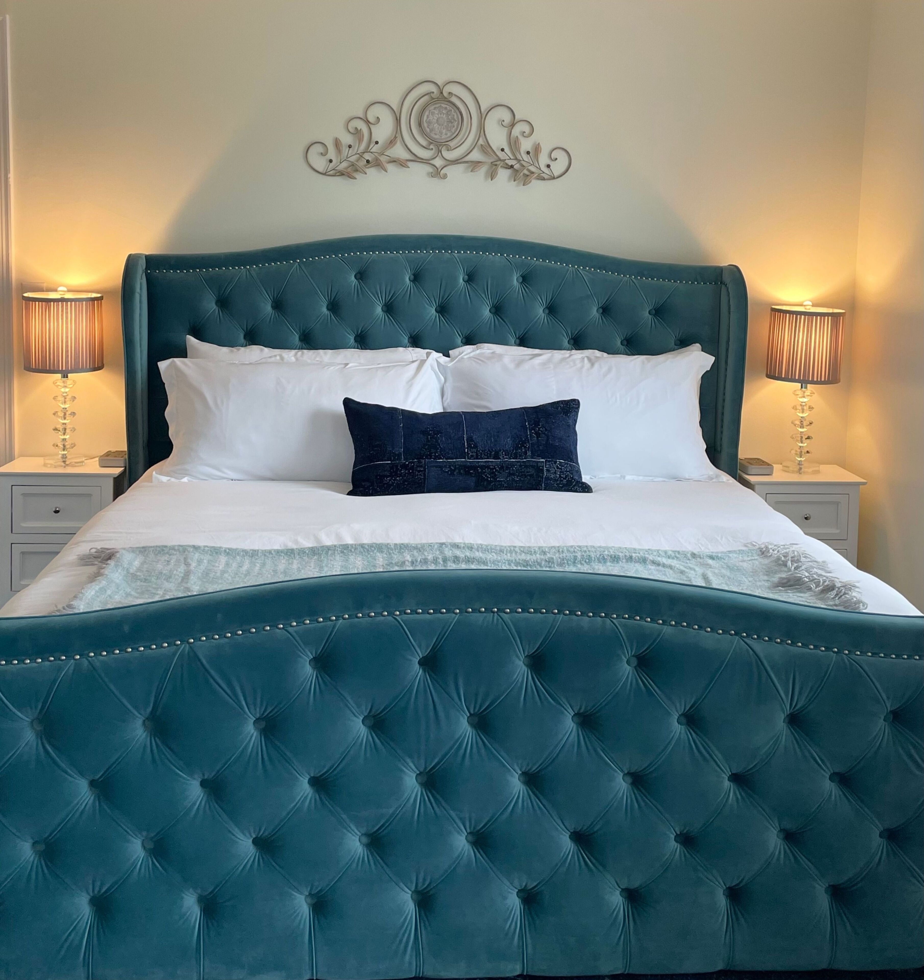 Een bed met een smaragdgroen bekleden hoofdeinde, witte en donkerblauwe kussens en een decoratief kunstwerk erboven.