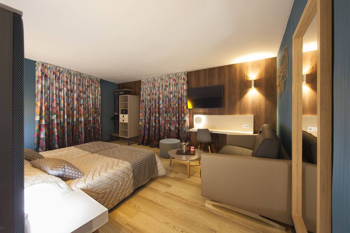 A modern hotel room with a bed, a sofa, a desk, and a TV.
