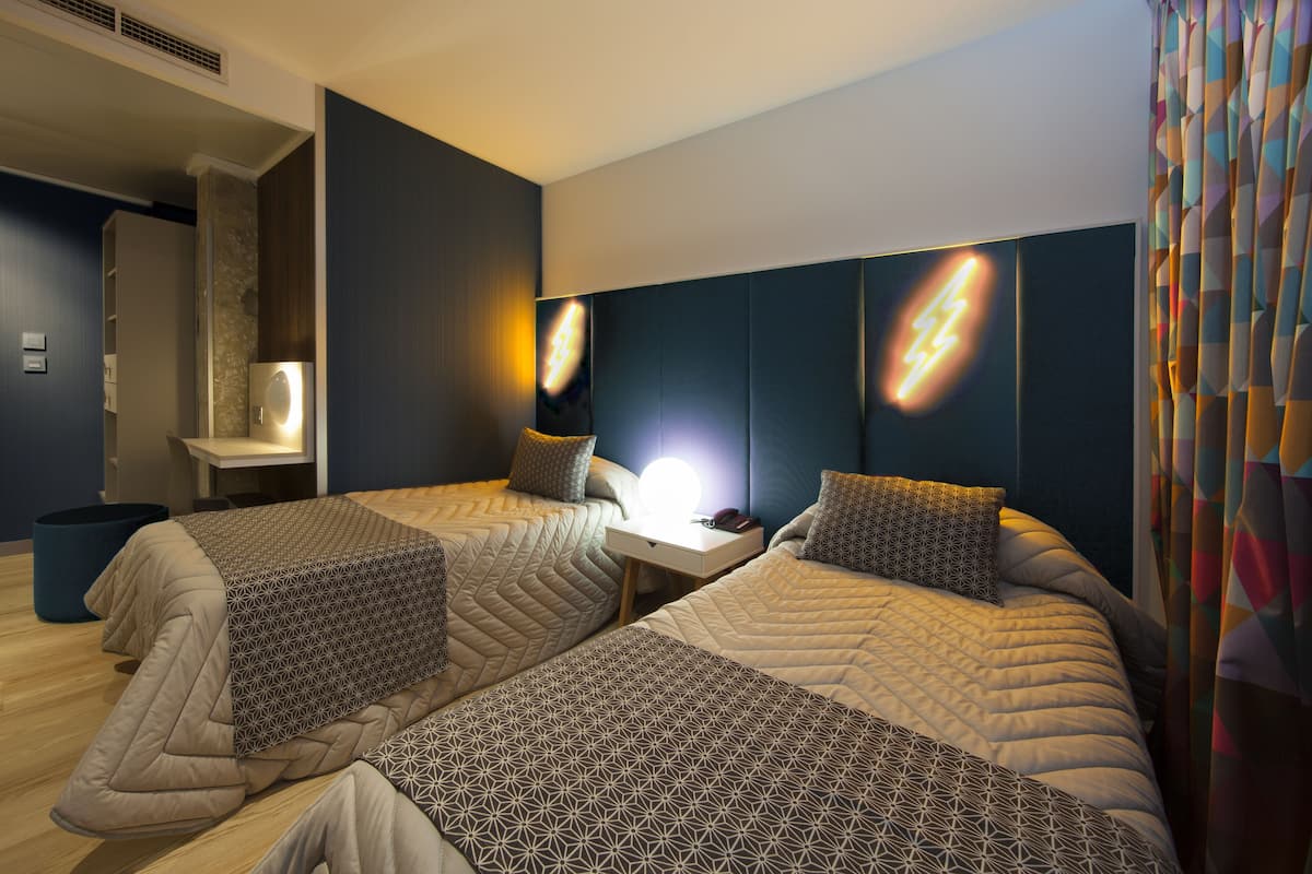 A modern hotel room with two beds, a bedside table with a lamp, and a wall-mounted light fixture.