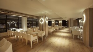 A modern interior with white chairs and tables arranged in rows, wooden floors, and circular light fixtures on the ceiling. - Hotel Cuco (Benidorm)