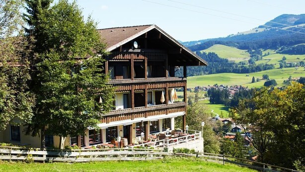 A two-story wooden building with a balcony, surrounded by lush greenery and a view of a valley. - Berghotel Maderhalm (Fischen im Allgaeu)