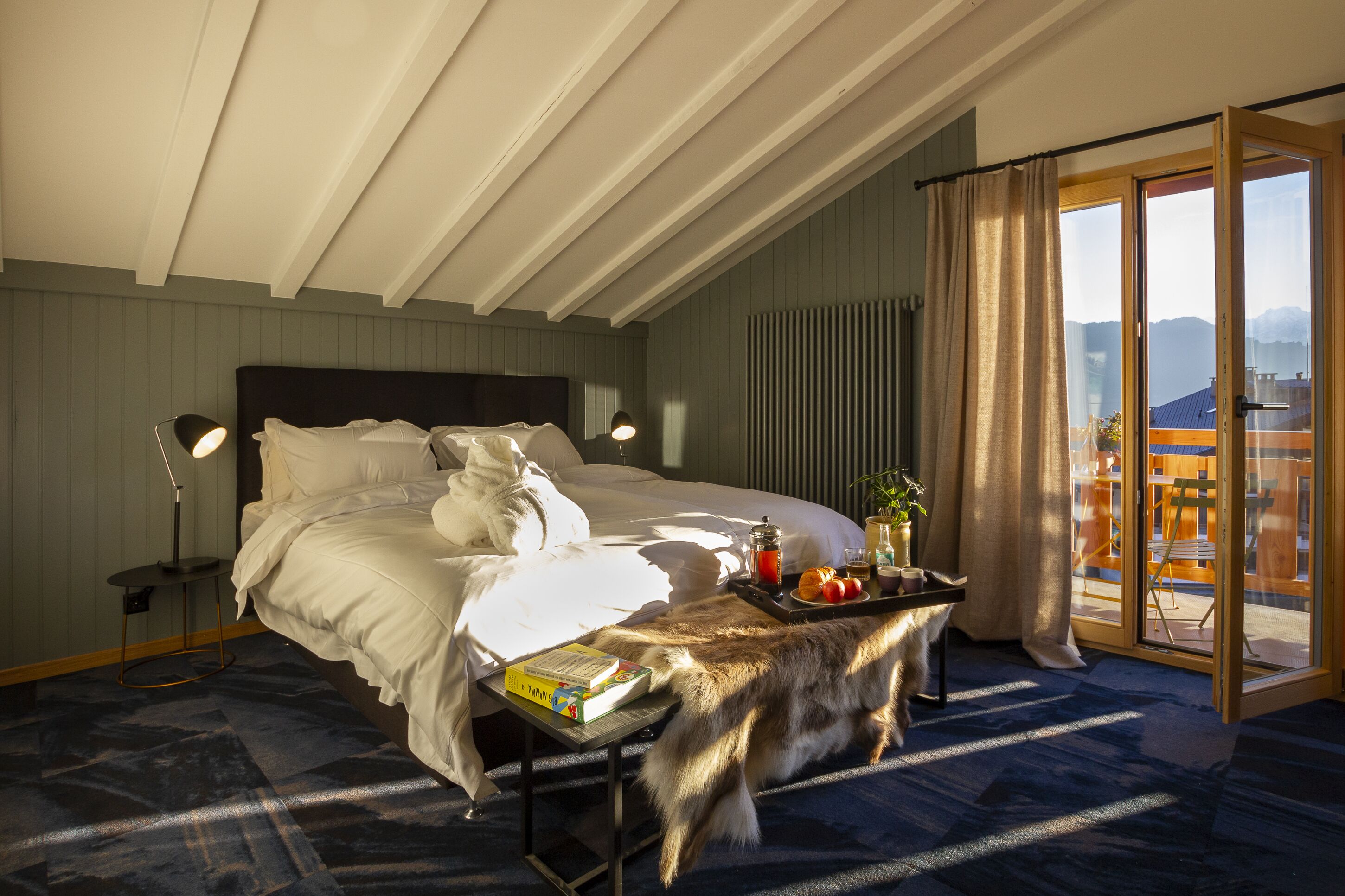A bedroom with a large bed, a bedside table with a book, a tray with fruits and a drink, and a view of the outdoors through a sliding door.
