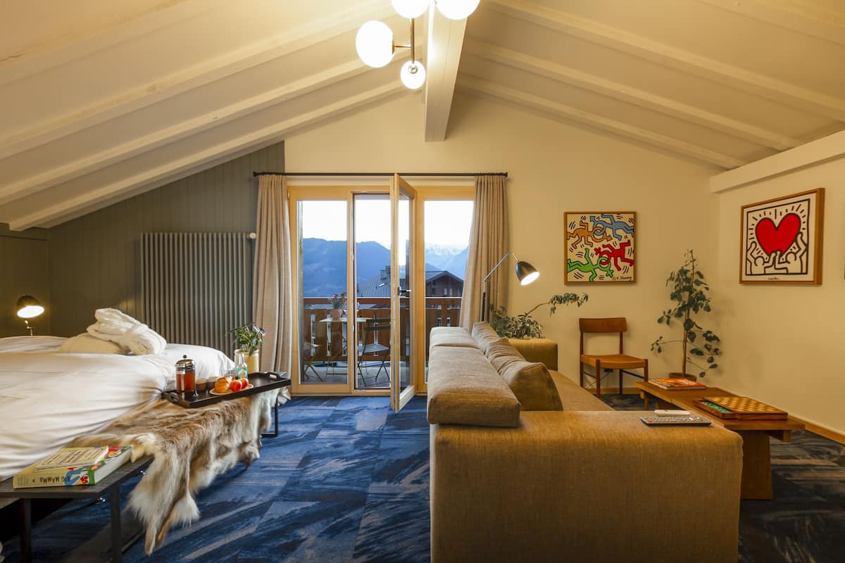 Ein Hotelzimmer mit Bett, einer Couch, einem Esstisch und Blick auf Berge durch die Fenster.