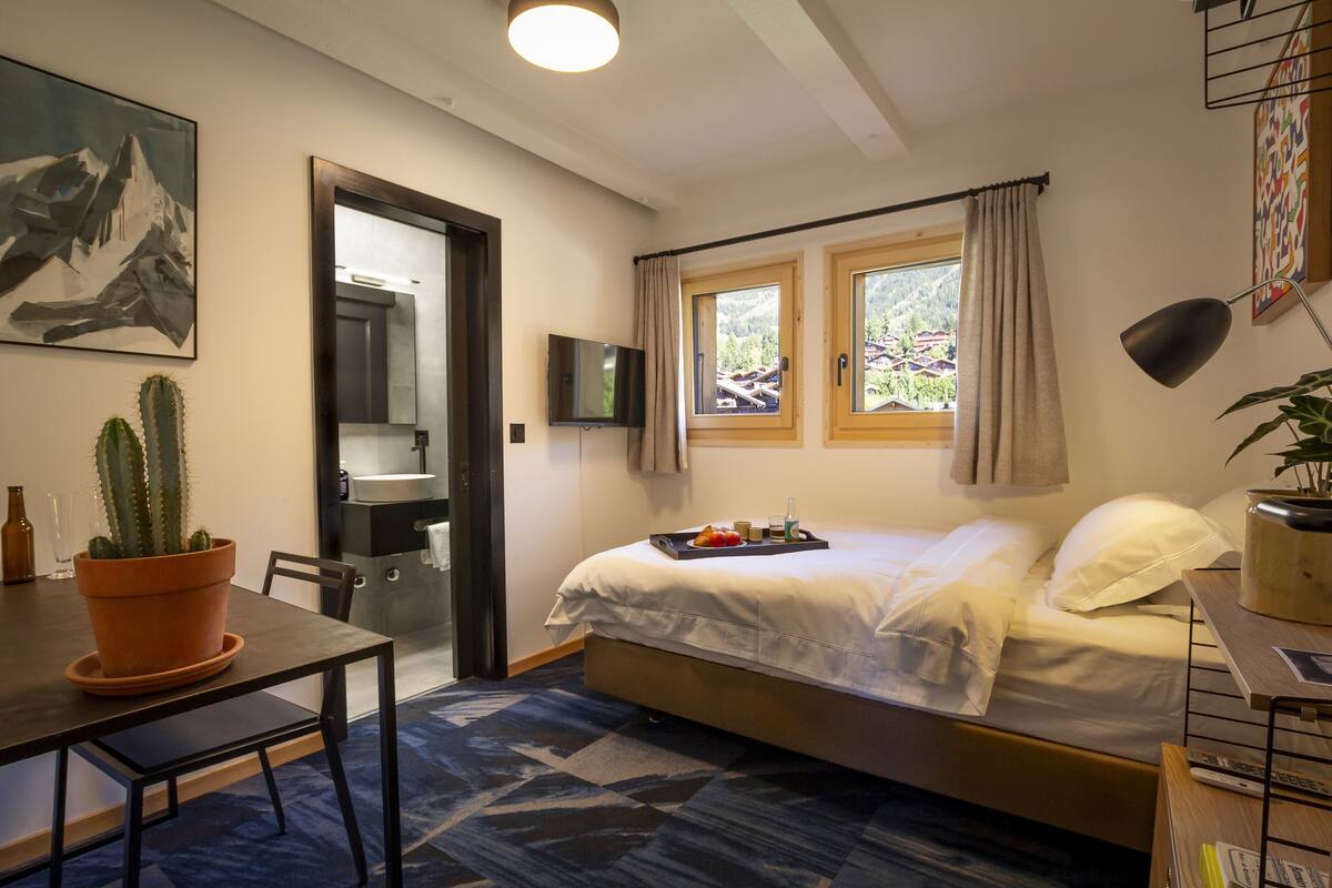Ein Schlafzimmer mit Bett, Schreibtisch, Zimmerpflanze, Fernseher und einem Fenster mit Vorhängen.