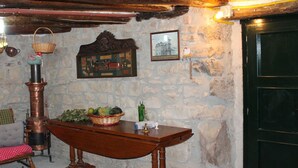 Una habitación rústica con pared de piedra, una mesa de madera con cesta de frutas, estufa de cobre y una puerta negra.